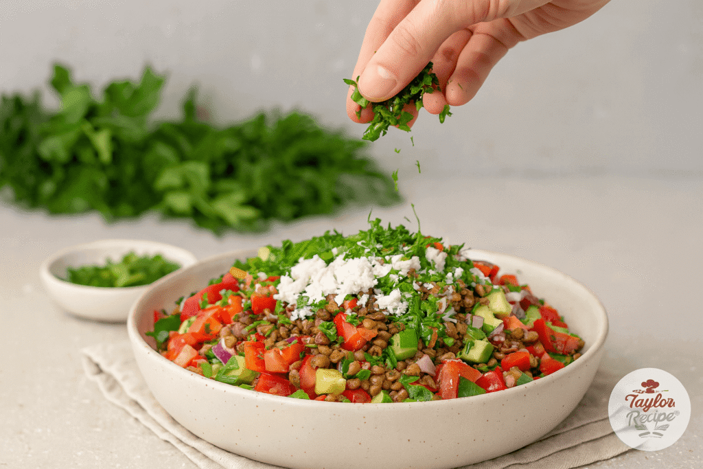 Mediterranean lentil salad in a bowl with colorful vegetables, topped with crumbled feta cheese and fresh herbs being sprinkled on top.